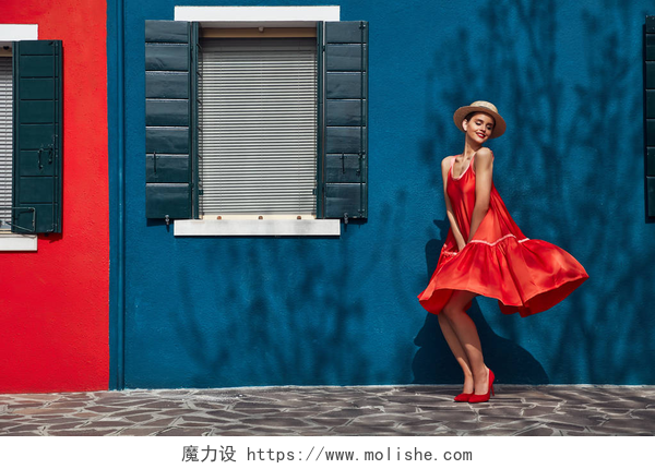 蓝色墙壁前的穿着红色裙子女人穿着红色连衣裙和高跟鞋的快乐年轻女子头上戴着草帽, 在布拉诺岛上摆着蓝色和红色的房子。墙上有树荫